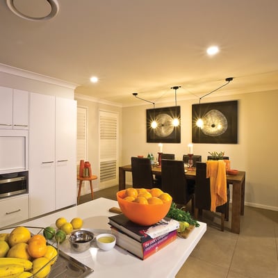 Et moderne køkkenalrum med spisebord og stole, belyst af loftslamper og moderne pendel lamper, med frugtskål på bordet.