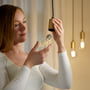 Kvinde installerer WiZ LED klar filament pære med E27 fatning i en lampeholder, med flere lignende pærer tændt i baggrunden.