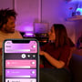 En mand og en kvinde taler sammen i en stue, mens en Philips Hue Go lampe lyser med lilla nuancer, og en smartphone viser Hue appen.