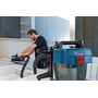 Bosch GAS 18V-10L Professional-støvsuger er koblet til slangen mens en håndverker arbeider med elektroverktøy ved en benk.